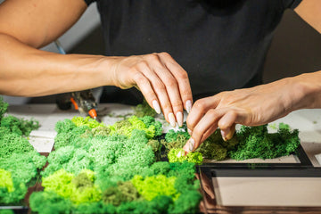 A woman seen from a different angle as she carefully places vibrant green moss into a picture frame. Her hands work delicately to create a textured, natural art piece, bringing an organic and earthy touch to the design.